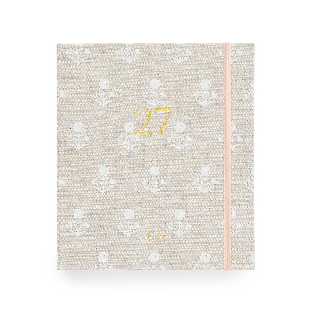 A 2027 fabric planner with a beige linen cover, featuring a white floral pattern, a gold foil '27' on the front, and a pale pink elastic closure band with "CS' in the bottom center
