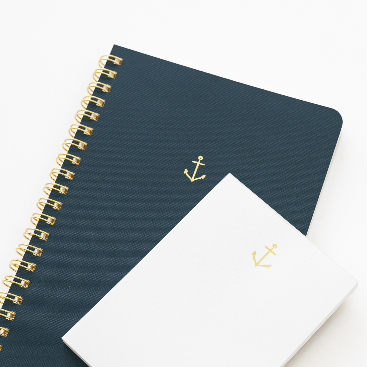 A navy blue spiral-bound notebook with a gold anchor, layered over a small white notepad with a matching gold anchor