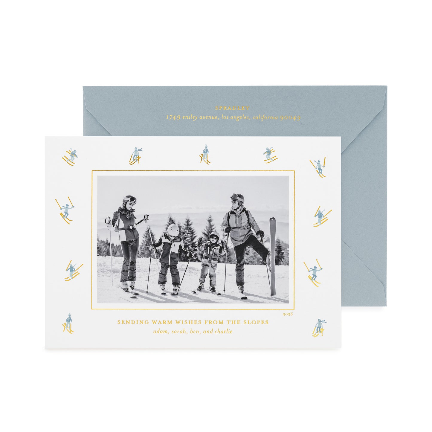 A personalized ski-themed family holiday photo card with a black-and-white family photo in the center, surrounded by a border of small blue and gold skiers, shown with a light blue envelope with gold foil details and the card printed with 'sending warm wishes from the slopes' 
