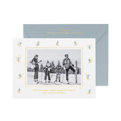 A personalized ski-themed family holiday photo card with a black-and-white family photo in the center, surrounded by a border of small blue and gold skiers, shown with a light blue envelope with gold foil details and the card printed with 'sending warm wishes from the slopes' 