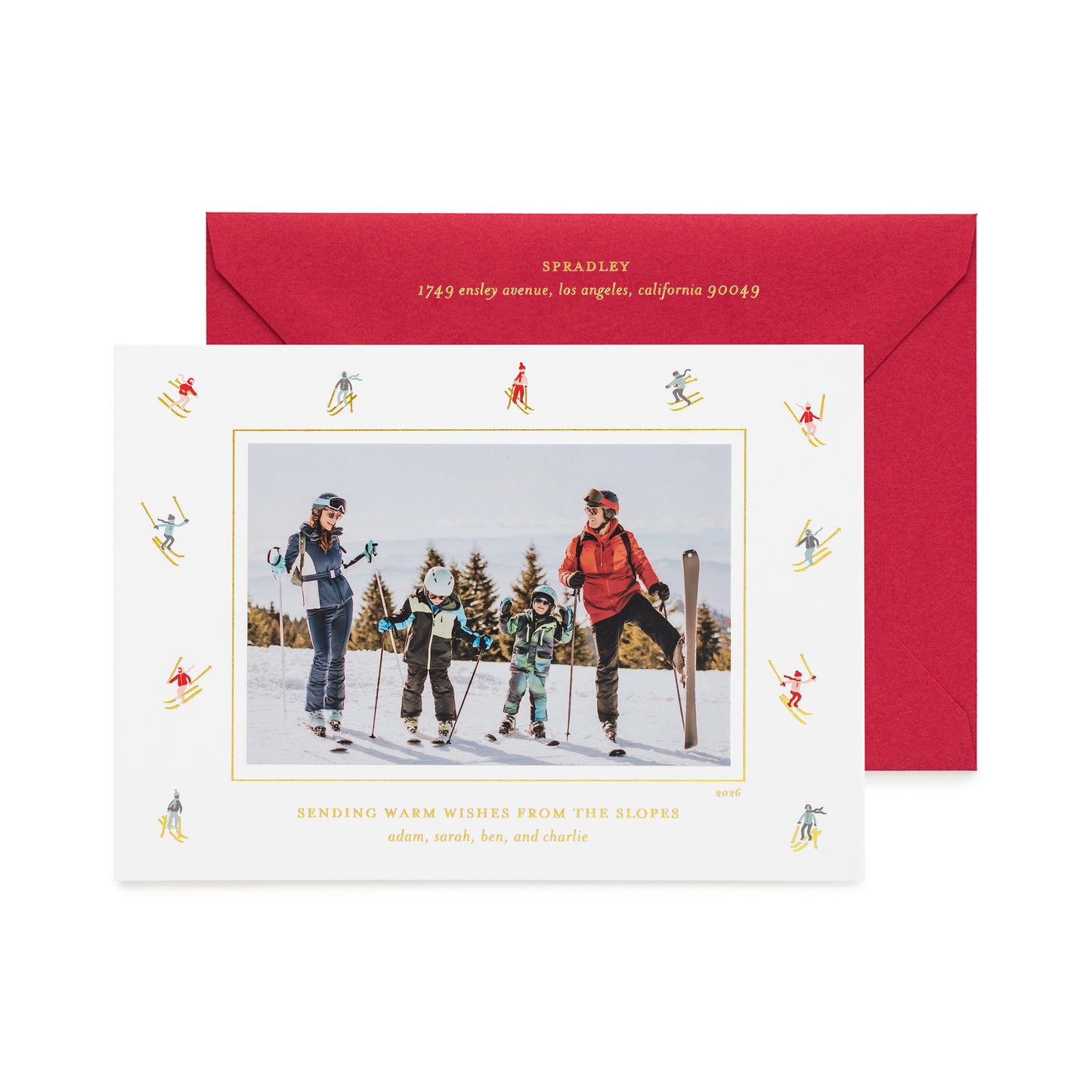 A personalized ski-themed holiday photo card with a black-and-white family photo in the center, surrounded by a border of small blue and gold skiers, shown with a red envelope with gold foil details and the card printed with 'sending warm wishes from the slopes' 