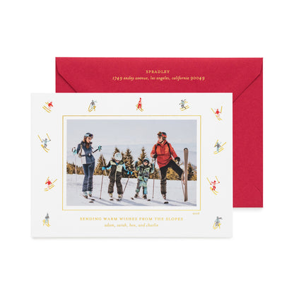A personalized ski-themed holiday photo card with a black-and-white family photo in the center, surrounded by a border of small blue and gold skiers, shown with a red envelope with gold foil details and the card printed with 'sending warm wishes from the slopes' 