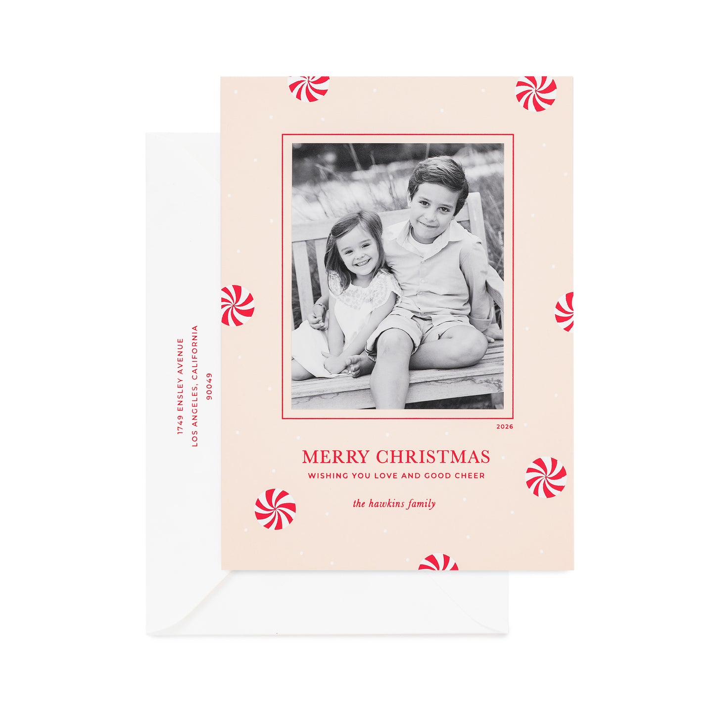 A personalized Christmas photo card on pale pink paper with, red and white peppermint candy icon pattern, a black-and-white family photo, printed with "merry christmas wishing you love and good cheer" with a white envelope printed the address in red ink