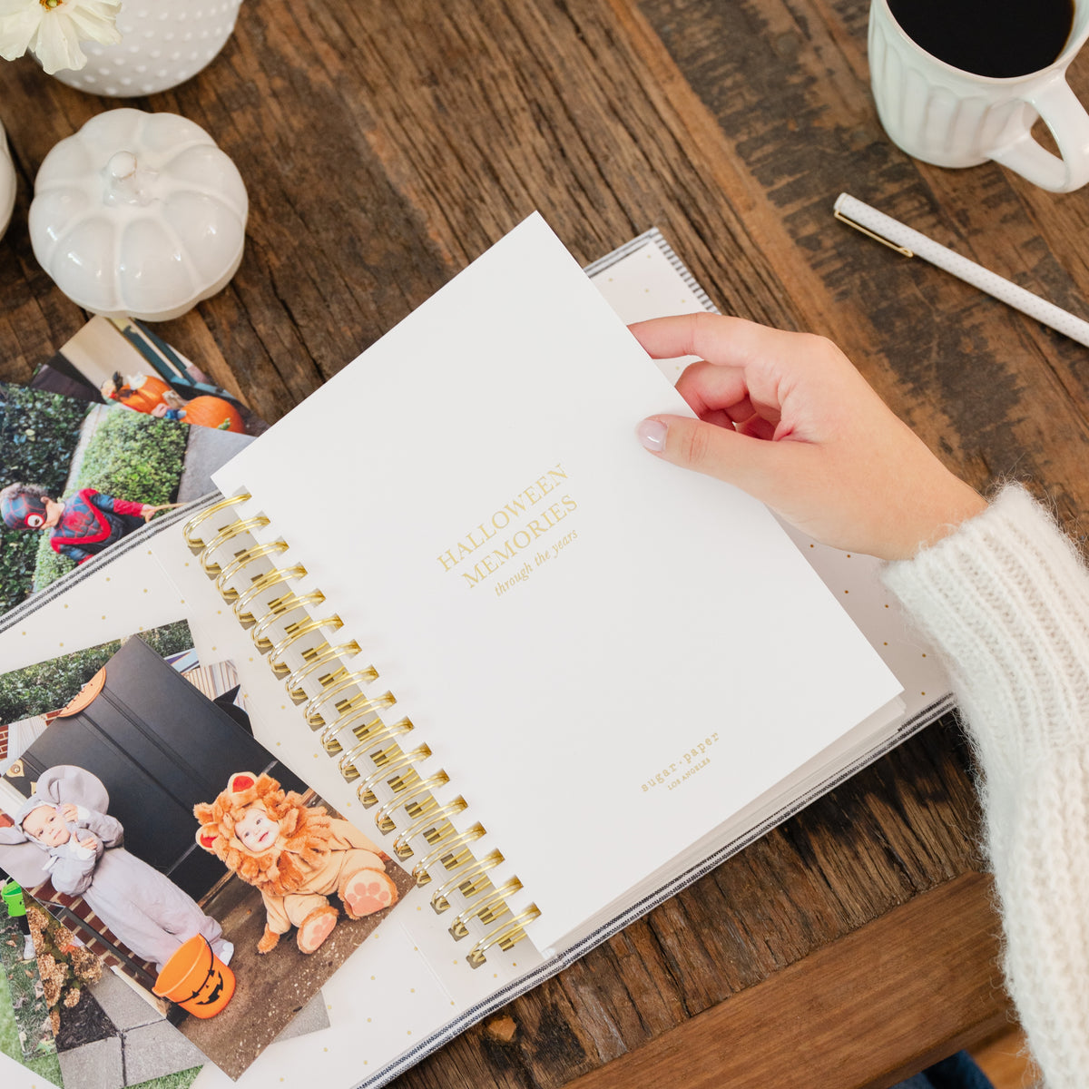 A hand wearing a white knit sweater holds an open white spiral-bound journal on a rustic wooden table. The cover reads 'HALLOWEEN MEMORIES' in gold lettering with photos of kids in halloween costumes and pumpkins in the background