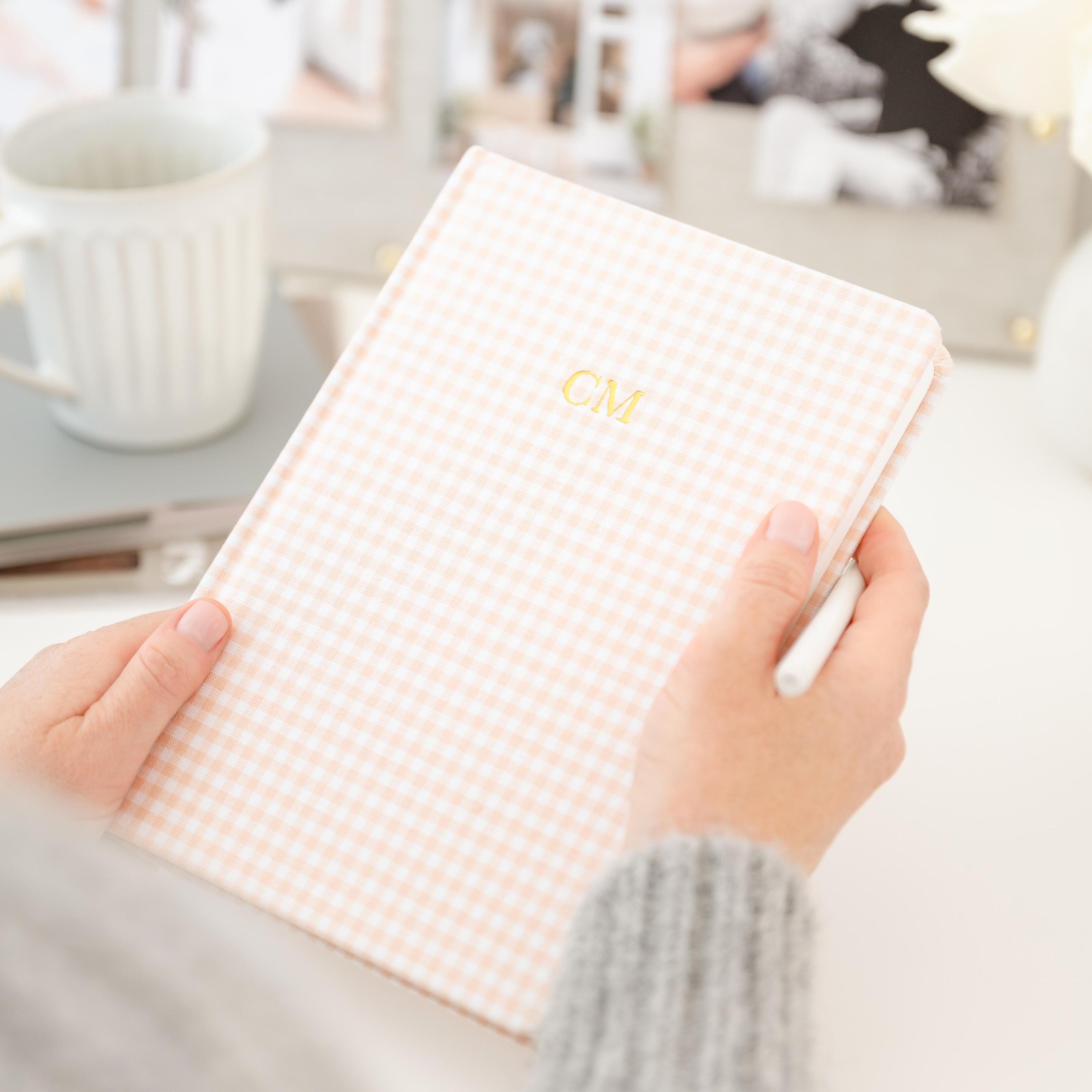 A person holding a light pink and white gingham check journal with gold foil initials 'CM' on the cover at a desk with coffee cup and inspiration board in background