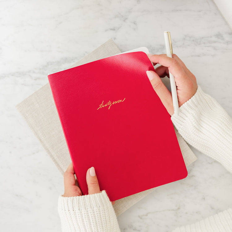 A top-down view of a person holding a bright red leather planner with gold cursive lettering with 'twenty seven' on a white marble desk. The person is wearing a white ribbed sweater and holding a white pen.