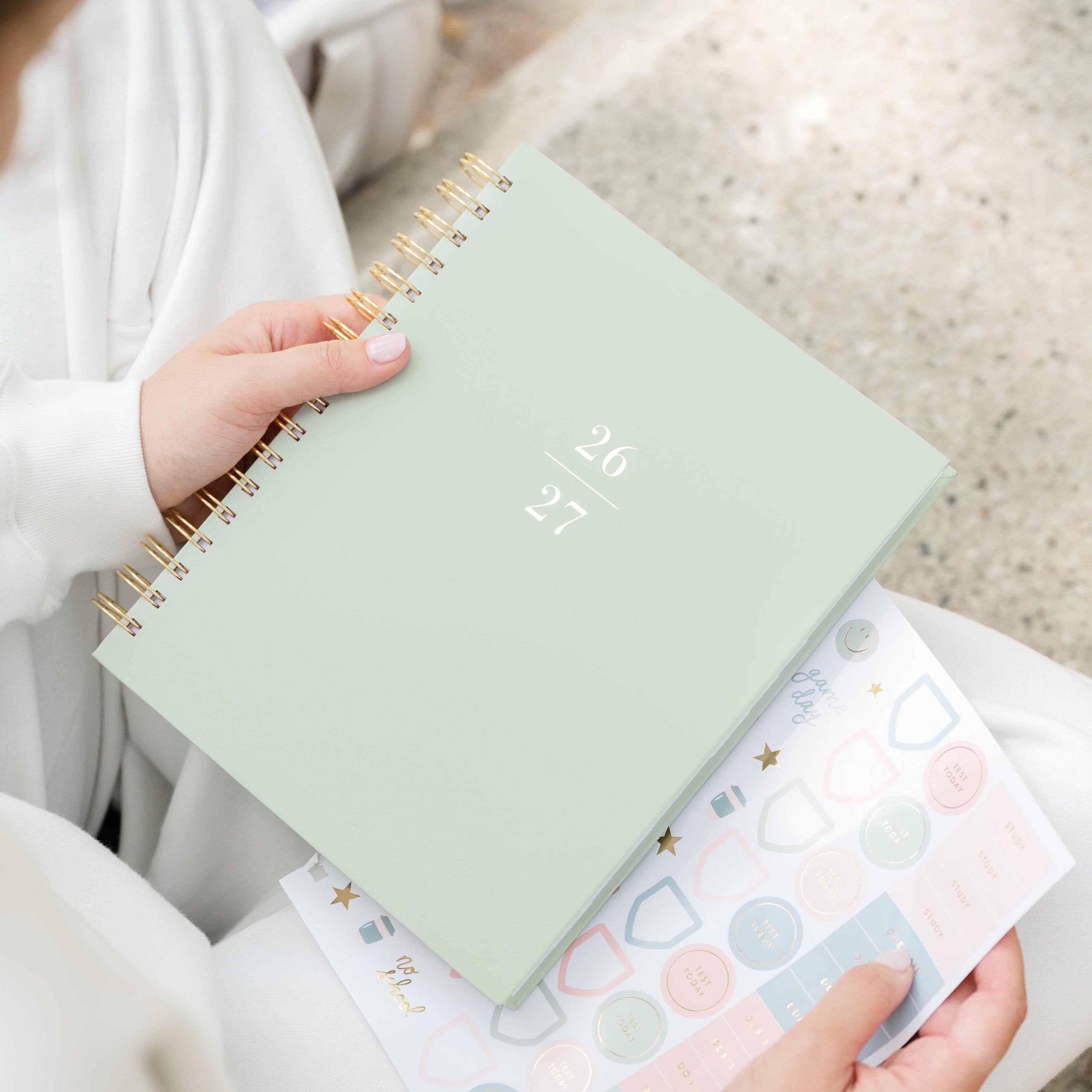 A person holds a mint spiral-bound planner with gold wire binding. The cover shows the dates '26' and '27' in gold foil with sheet of stickers in hand