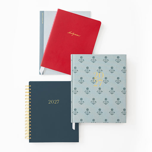 Four planners arranged on a white background: a red hardcover with gold script, a dark navy spiral-bound 2027 planner, a light blue floral patterned hardcover with gold foil '2027' and a blue and white striped planner